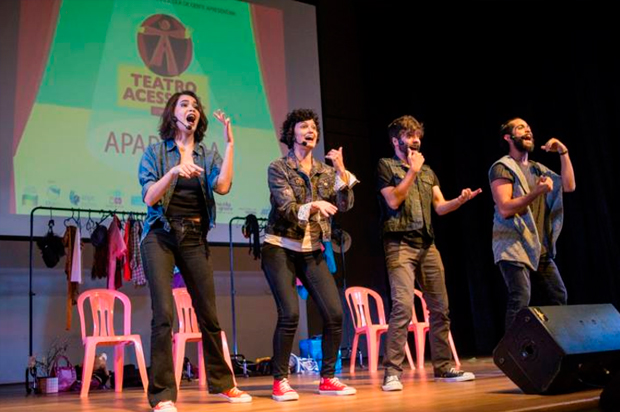 Duas atrizes e dois atores estão no palco, lado a lado, dançando e ao mesmo tempo fazendo Libras com a letra da música. Eles/as usam microfone de lapela, levantam os braços como se estivesse convidando a plateia a se aproximarem. No palco, quatro cadeiras, duas araras com roupas penduradas, acessórios no chão..No telão, logo atrás, a logo do projeto no telão.