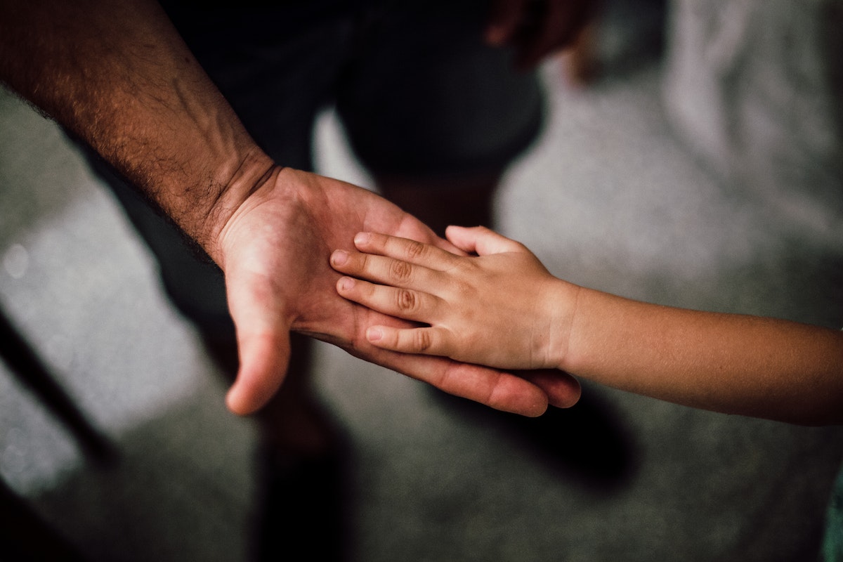 Sobre fundo desfocado de um card retangular, foto em primeiro plano mostra uma mão masculina estendida, apoiando com a palma a mãozinha de uma criança; os braços de ambas aparecem parcialmente.