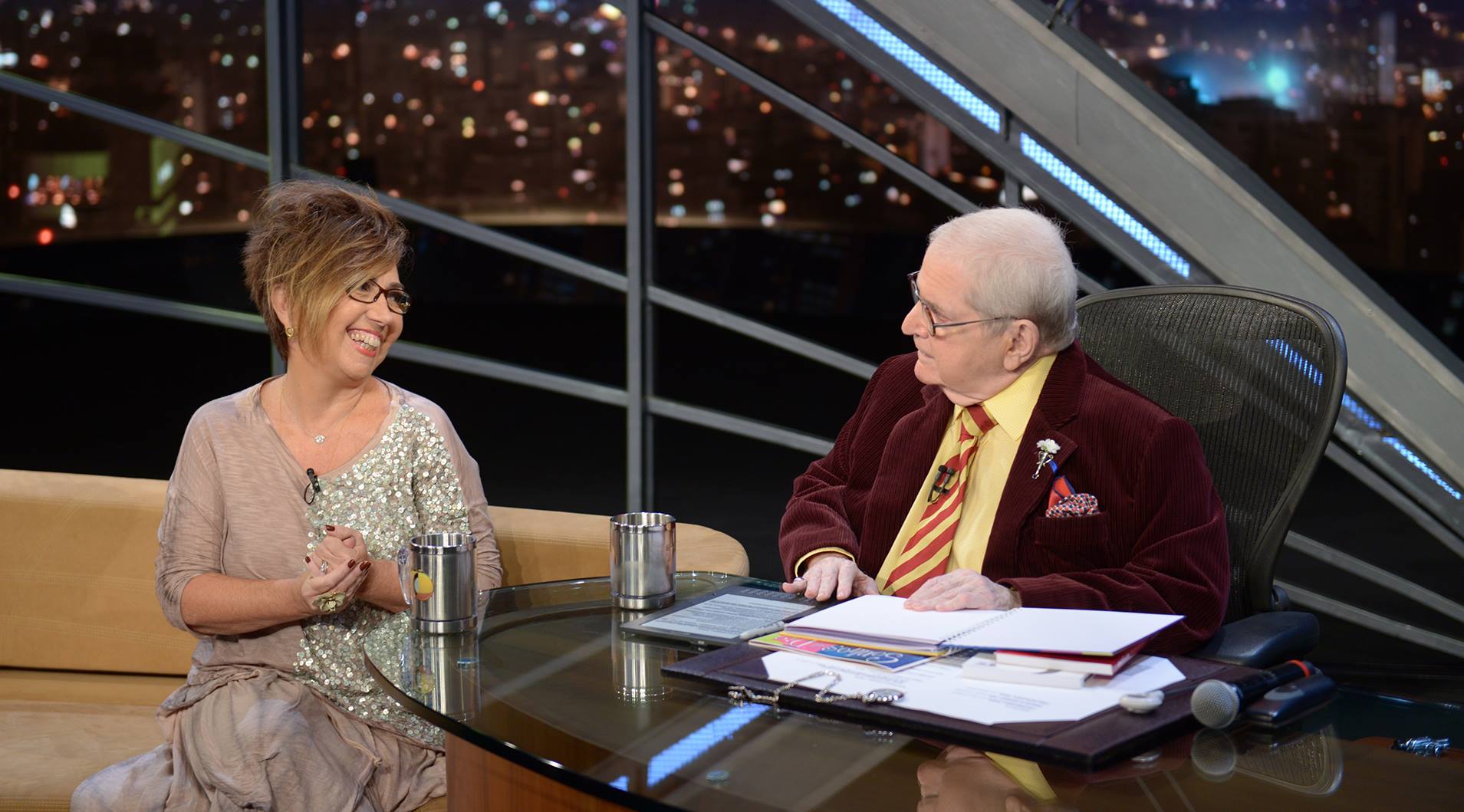 Foto mostrando Claudia Werneck, sentada no sofá, sorridente, conversando com o apresentador Jô Soares também sentado na sua mesa.