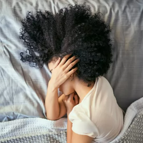 A fotografia mostra, de cima, uma mulher negra deitada de lado em uma cama. Ela tem cabelos crespos, que aparecem repousados em forma de leque sobre o travesseiro; está cobrindo o rosto com uma das mãos e, com a outra, apoia o queixo. Ela está coberta até a altura dos ombros com um cobertor que, assim como a roupa de cama, é de cor cinza. 