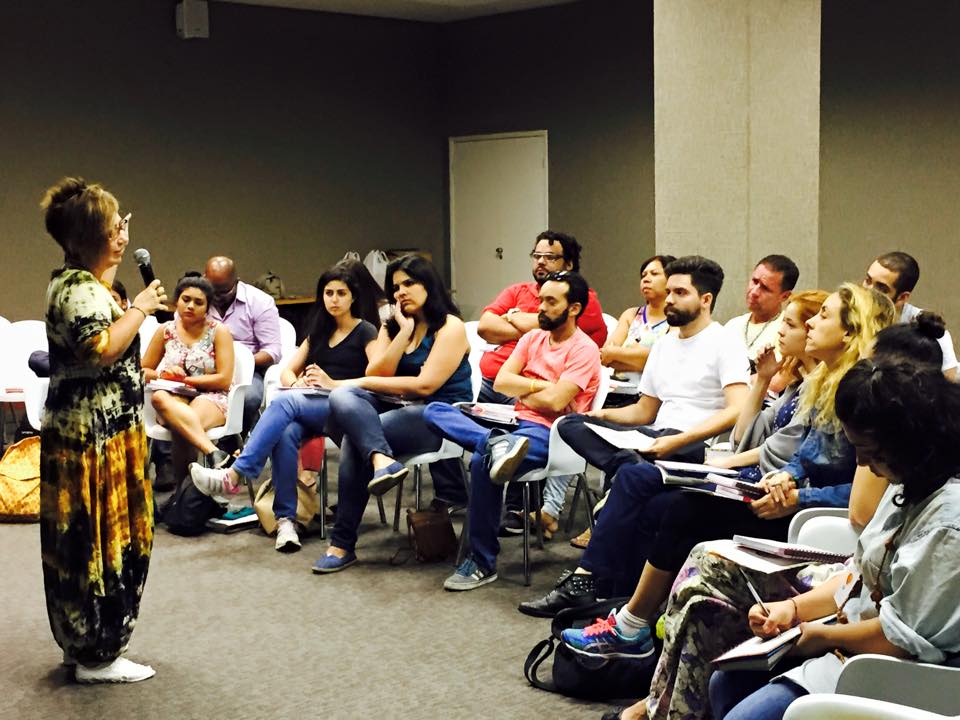 Foto mostrando Cláudia Werneck, de pé, falando ao microfone com turma de dezenas de alunos, sentados, prestando atenção.