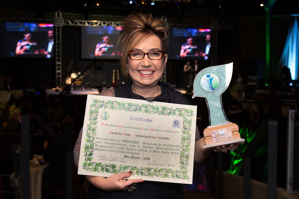 Claudia Werneck segura com a mão direita o certificado e com a mão esquerda o troféu do "Prêmio Socioambiental Chico Mendes". Ela está sorridente. Atrás dela estão vários telões com imagens desfocadas.