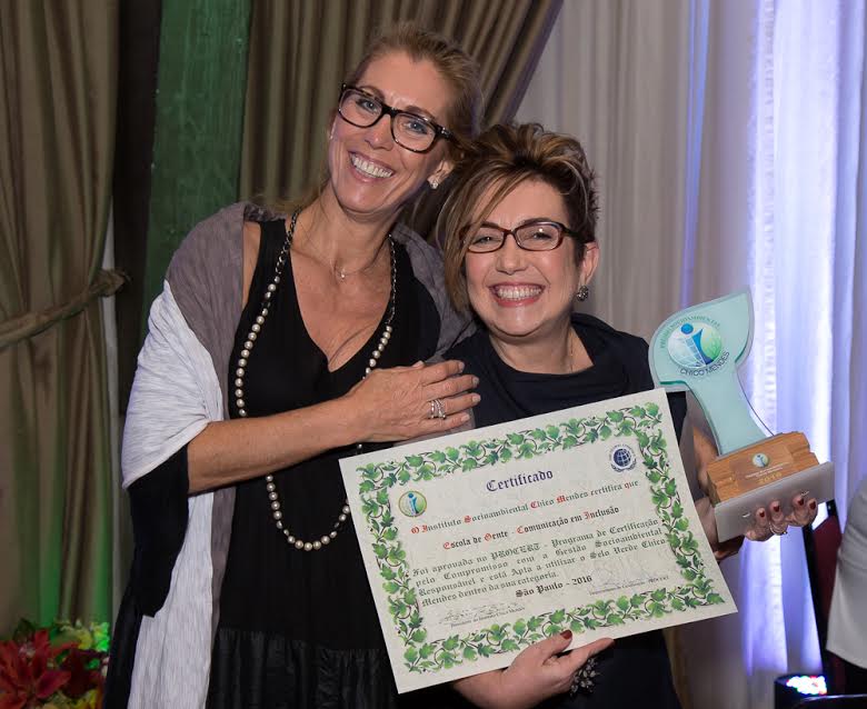 Rosana Fonseca, conselheira fiscal da Escola de Gente, e Claudia Werneck estão abraçadas, lado a lado, em pé. Claudia segura o certificado e o troféu.