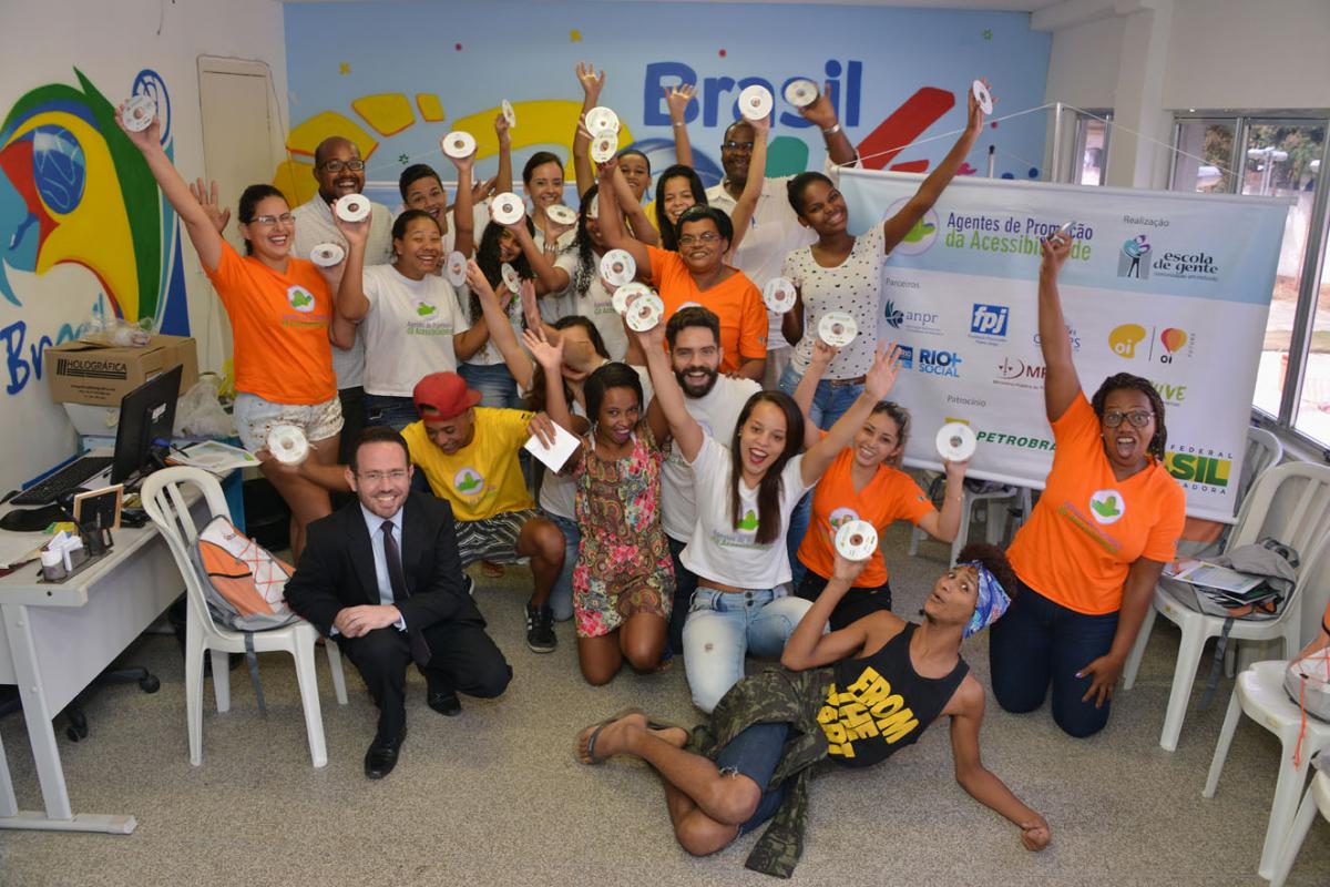 Foto mostrando várias pessoas, entre homens e mulheres, em pé e sentados/as, posam para a foto levantando os braços, segurando os Cd’s do Livro Falado. Atrás deles, no canto direito, um banner com a barra de logos dos parceiros e patrocinadores do projeto “Agentes de Promoção da Acessibilidade”.