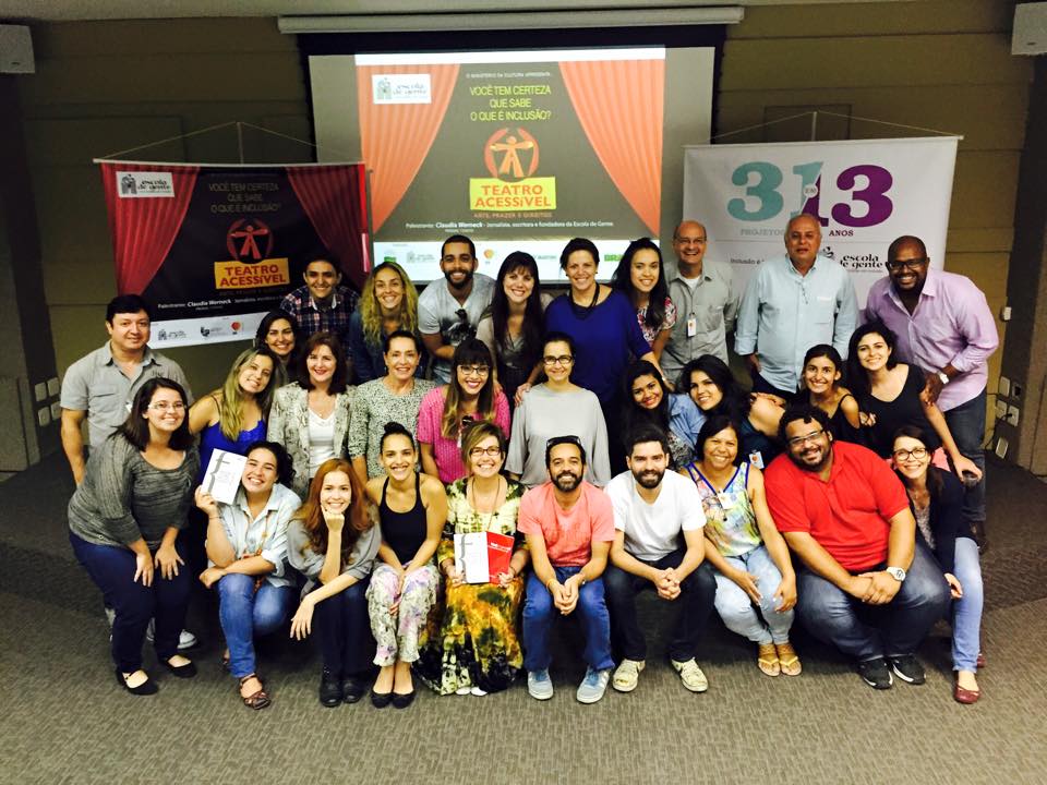 Foto mostrando a turma que participou da palestra, todos olhando para a câmera sorridentes, com o cartaz do Teatro Acessível ao fundo.