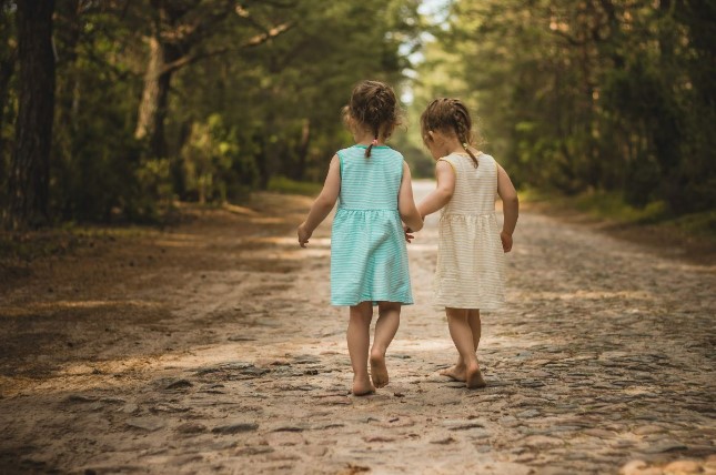 Fotografia retangular horizontal traz foto a cores de uma estrada com chão coberto de folhas, onde centralmente caminham de mãos dadas duas meninas em torno de 4 anos. Elas estão de costas para a câmera, descalças e ambas têm o cabelo castanho claro preso ao alto em uma trancinha que cai na nuca; a da esquerda usa vestido azul e a da direita branco. As laterais do caminho estão cobertas de árvores, com muito verde.