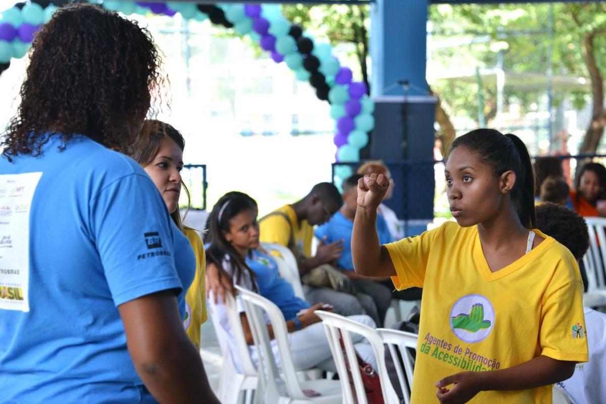 Fotografia colorida horizontal. Uma jovem sinaliza em Libras, para uma mulher. No fundo cadeiras de plástico brancas, com algumas pessoas sentadas. A jovem está à direita, ela é negra, cabelo preso num rabo de cavalo, olhos castanhos, usa blusa amarela com estampa escrita Agentes de Promoção de Acessibilidade. Ela está com a mão direita erguida, com todos os dedos fechados. À esquerda, a mulher está de frente para a jovem e de costas para nós. Ela é negra, cabelos cacheados, castanhos, na altura do ombro, usa camiseta azul. Ao lado dela aparece o rosto de outra mulher, de pele clara, olhos castanhos, que olha para a jovem.