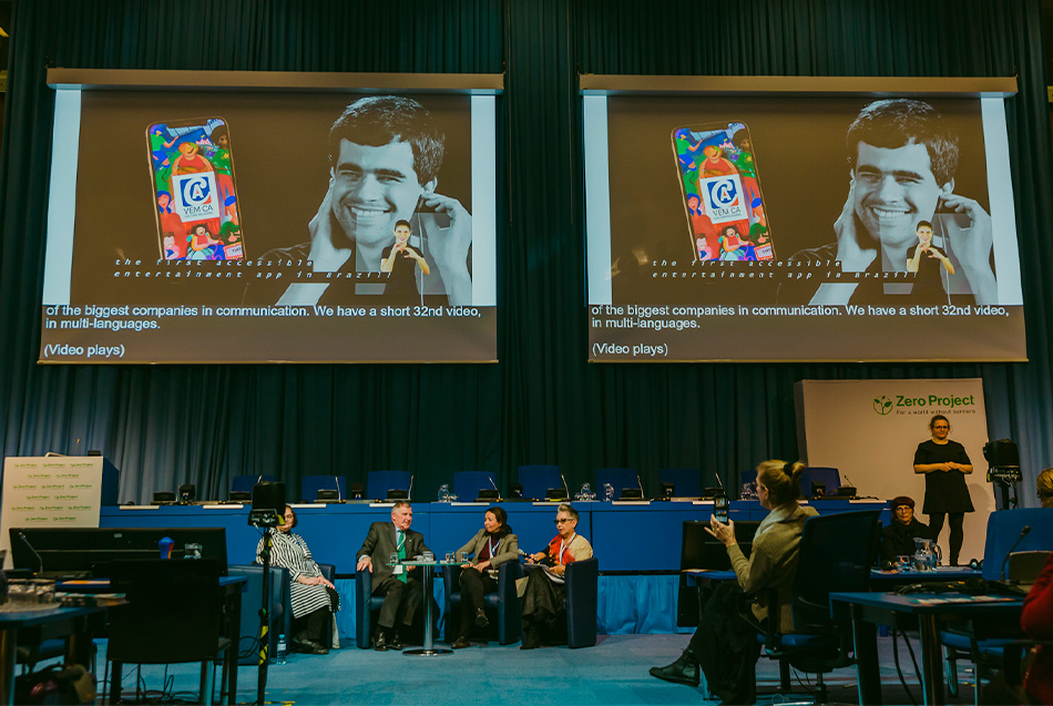 Fotografia colorida, Claudia Werneck e outras três pessoas, sentadas em poltronas, no chão da plateia, na frente de um palco. Penduradas, sobre o palco, duas telas mostram o aplicativo VEM CA, à direita a intérprete de Libras.