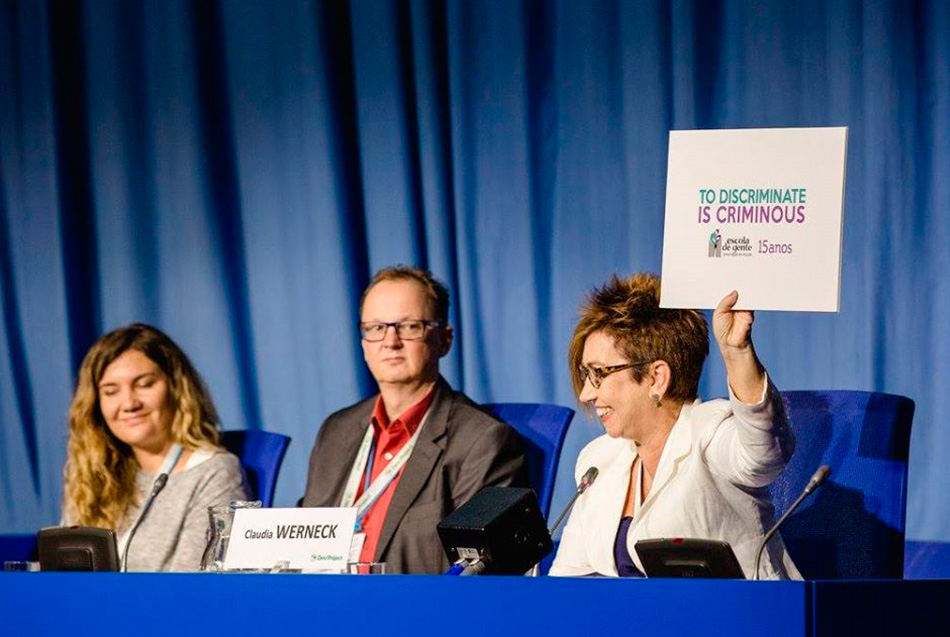 Fotografia colorida, Claudia Werneck, numa bancada ao lado de uma mulher e um homem, segura cartaz "To discriminate is criminous" no lançamento desta campanha durante a Zero Conference.