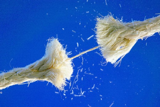 Sobre retângulo com fundo azul, foto enfoca uma corda de sisal esticada e rebentada em dois pedaços, unida apenas por um fio central. No seu entorno, vários resíduos da corda flutuam contra o azul.