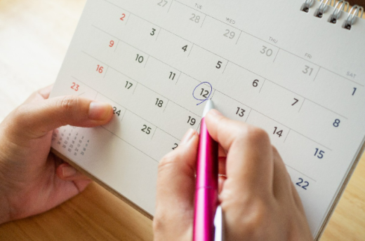 Sobre um retângulo, fotografia traz duas mãos com um calendário espiralado, que aparece com cinco linhas de numerais referentes aos dias de um mês. A mão direita segura uma caneta na cor rosa, que está marcando com um círculo preto o dia 12 neste calendário.