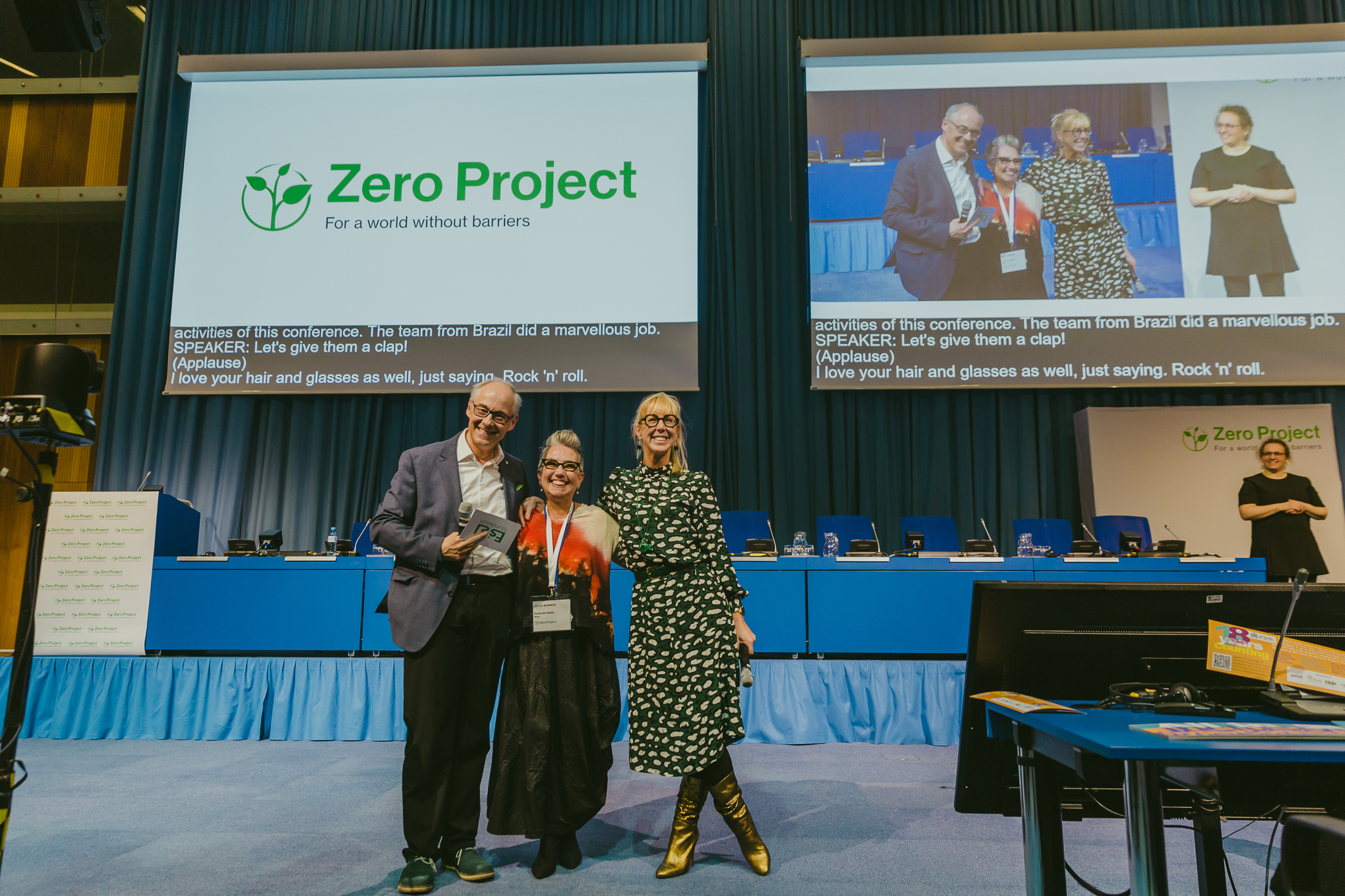 Fotografia colorida, Claudia Werneck, entre um homem e uma mulher, sorriem. Estão na frente do palco da Zero Conference.