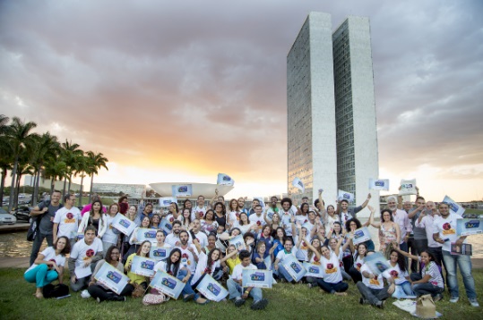 Fotografia colorida, grupo de pessoas de várias idades, gêneros e cores, usam camisa amarela. Todas sorrindo, posam em volta do banner do projeto.