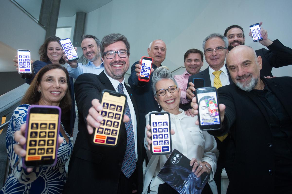 Fotografia colorida, Claudia Werneck e um grupo de pessoas, sorriem, e seguram celulares com a tela aberta no aplicativo VEM CA