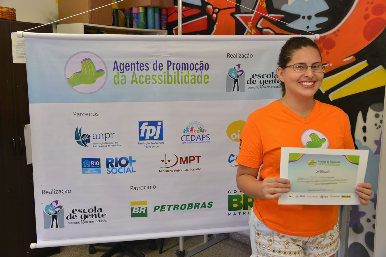 Descrição da Imagem: Marquênia de Oliveira Barbosa Silva, em pé, segurando o certificado de conclusão do projeto “Agentes de Promoção da Acessibilidade”, sorri posando para foto. Atrás dela, um banner com a barra de logos dos parceiros e patrocinador.