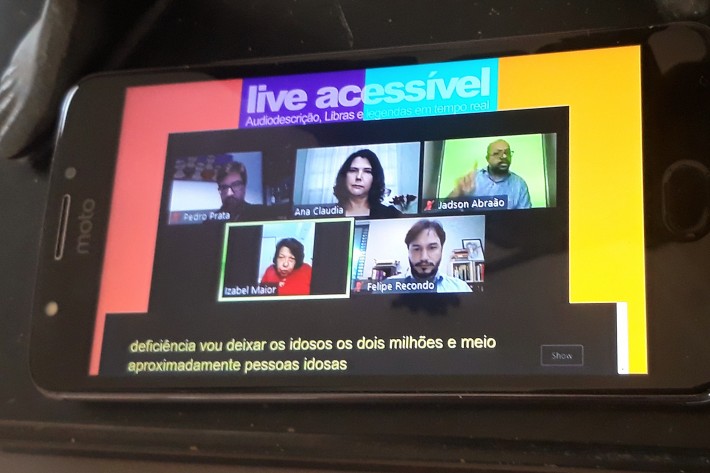 Em um smartphone, a transmissão de uma live acessível pela ONG Escola de Gente, com a tela dividida em cinco janelas com as imagens dos participantes, entre eles um intérprete de Libras e, abaixo, a legenda em tempo real.  