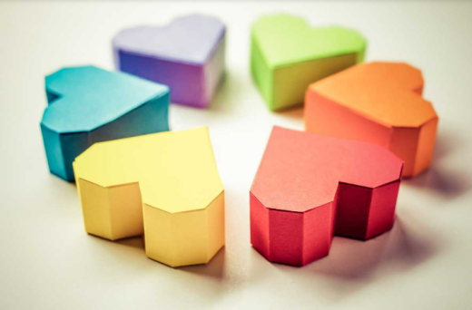 A imagem mostra 6 dobraduras de pequenas caixas em formato de coração, organizadas de modo a lembrar um trevo de quatro folhas. Cada caixa tem uma única cor, e as cores que aparecem são: amarelo, azul, roxo, verde, laranja e vermelho. Elas estão sobre uma superfície clara e reta.