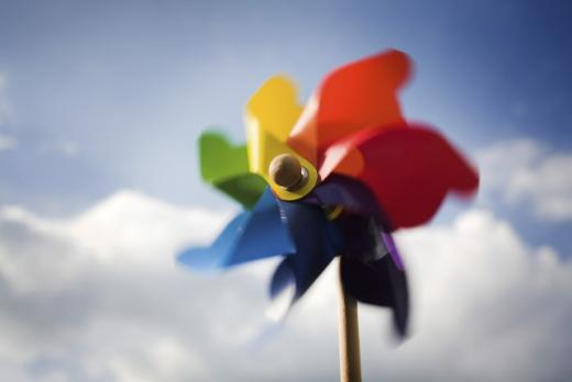 Sobre fundo de céu azul claro com nuvens brancas, em card retangular horizontal, um catavento multicolorido se destaca em primeiríssimo plano. Ele tem sete pás nas cores verde, amarela, laranja, vermelha, roxa, cinza e azul.